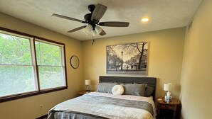 Une chambre à coucher avec un lit, un ventilateur de plafond, deux lampes de chevet, une fenêtre avec des stores et un tableau représentant un paysage urbain accroché au mur.
