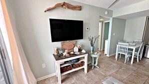 Un salon doté d’un téléviseur à écran plat, d’un meuble de rangement en bois, d’un coin repas avec des chaises blanches et d’un ventilateur de plafond.