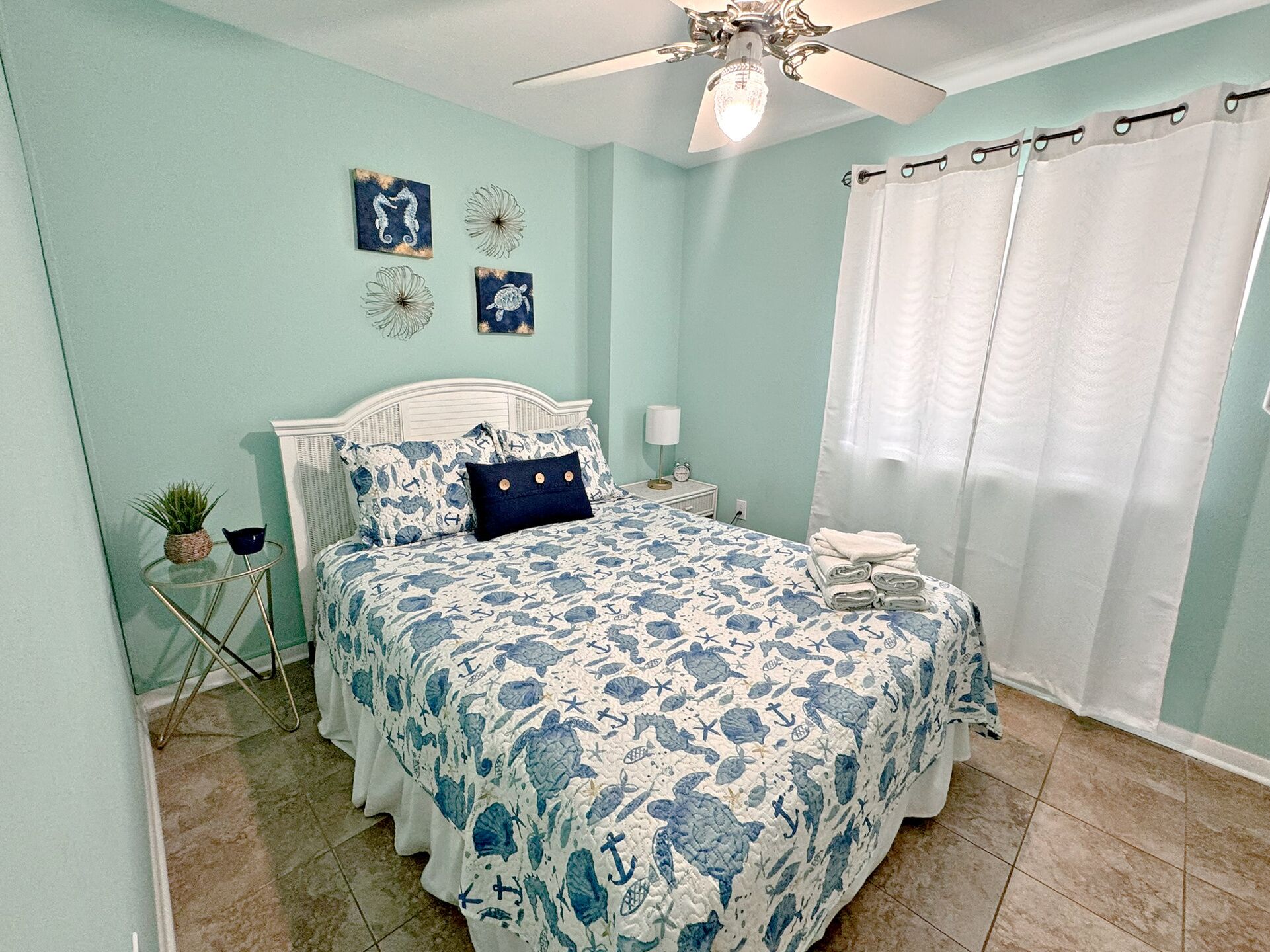 A bedroom with a bed, a ceiling fan, a nightstand, a lamp, a potted plant, and a window with curtains.