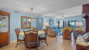 A spacious living room with a dining area, featuring a chandelier, wicker furniture, and a large window offering a view of the outdoors. - Castle Reef 213 stunning ocean views (New Smyrna Beach)