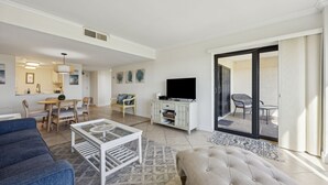 Una sala de estar moderna con un sofá, una mesa de centro y un televisor. Se aprecia un comedor con una mesa y sillas, y una cocina al fondo.