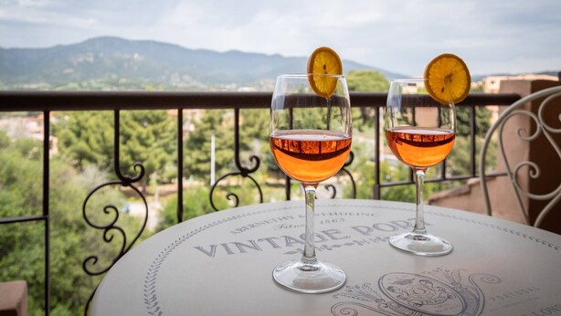 Deux verres d’une boisson ambrée sont posés sur une table au design rétro, le tout sur fond d’un paysage montagneux pittoresque.