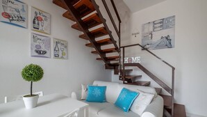 Un salon moderne avec un canapé blanc, une petite table et une plante en pot. On peut voir un escalier en bois sur lequel est accroché un panneau indiquant « HOME ».