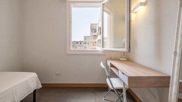 A small, bright room with a single bed, a desk, and a window overlooking a cityscape.