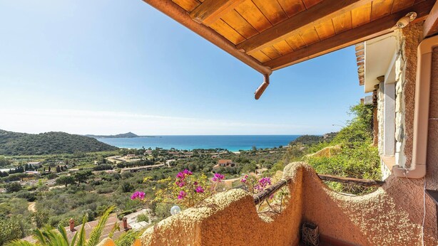 Vista sulla costa da un edificio rustico con tetto in legno, che si affaccia su un paesaggio pittoresco con spiaggia e colline.