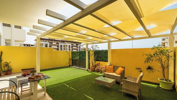 Un patio al aire libre con césped artificial, paredes amarillas y una pérgola que da sombra.