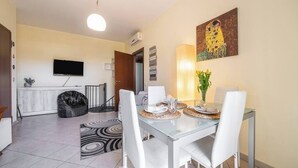 Una sala de estar moderna con una mesa de comedor de vidrio, sillas blancas, un sillón orejero negro y un televisor de pantalla plana montado en la pared, con un cuadro sobre la mesa.