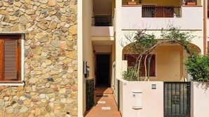 A narrow alleyway with a stone wall on one side and a building with a balcony on the other. There is a small garden area with a tree and a potted plant. - Relax Sea Home by Propertize - Villasimius (Villasimius)