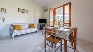 A dining area with a table set for four, a sofa with a patterned cover, and a television in the background. - Relax Sea Home by Propertize - Villasimius (Villasimius)
