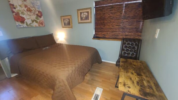 A bedroom with a bed, a small table, a wall-mounted TV, and a window with blinds.