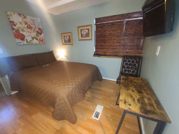 Un dormitorio con cama, una mesita, televisor montado en la pared y ventana con persianas.