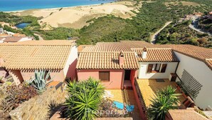 A residential area with terracotta-tiled roofs, a courtyard with potted plants, and a view of a beach and hills in the background.