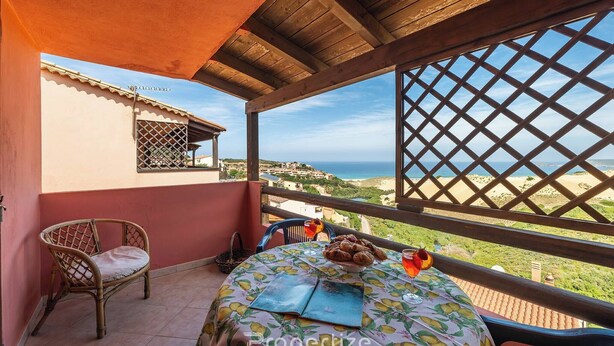 A balcony with a wooden pergola, a wicker chair, a table with a tablecloth, and a view of the sea and buildings.