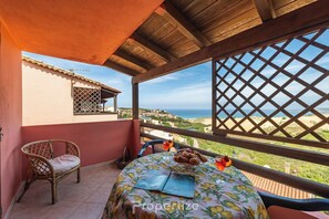 Ein Balkon mit einer Holz-Pergola, einem Korbstuhl, einem Tisch mit Tischdecke und Blick auf das Meer und Gebäude.