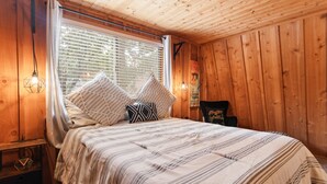 A cozy wooden room with a bed, a nightstand, and a window with blinds.
