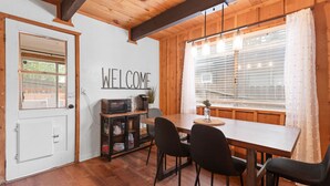 A wooden interior with a 'WELCOME' sign, a dining table with chairs, a coffee machine, and a window with blinds.