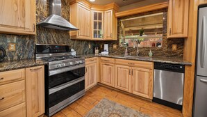A kitchen with wooden cabinets, granite countertops, stainless steel appliances, and a window overlooking a garden. - Moonridge Retreat - 6BR, Walk to Bear Mountain, Foosball Table, BBQ (Big Bear Lake)