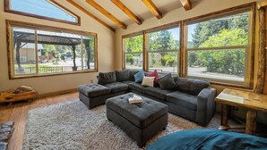 A spacious living room with a sectional sofa, ottoman, and large windows offering views of the outdoors. - Moonridge Retreat - 6BR, Walk to Bear Mountain, Foosball Table, BBQ (Big Bear Lake)