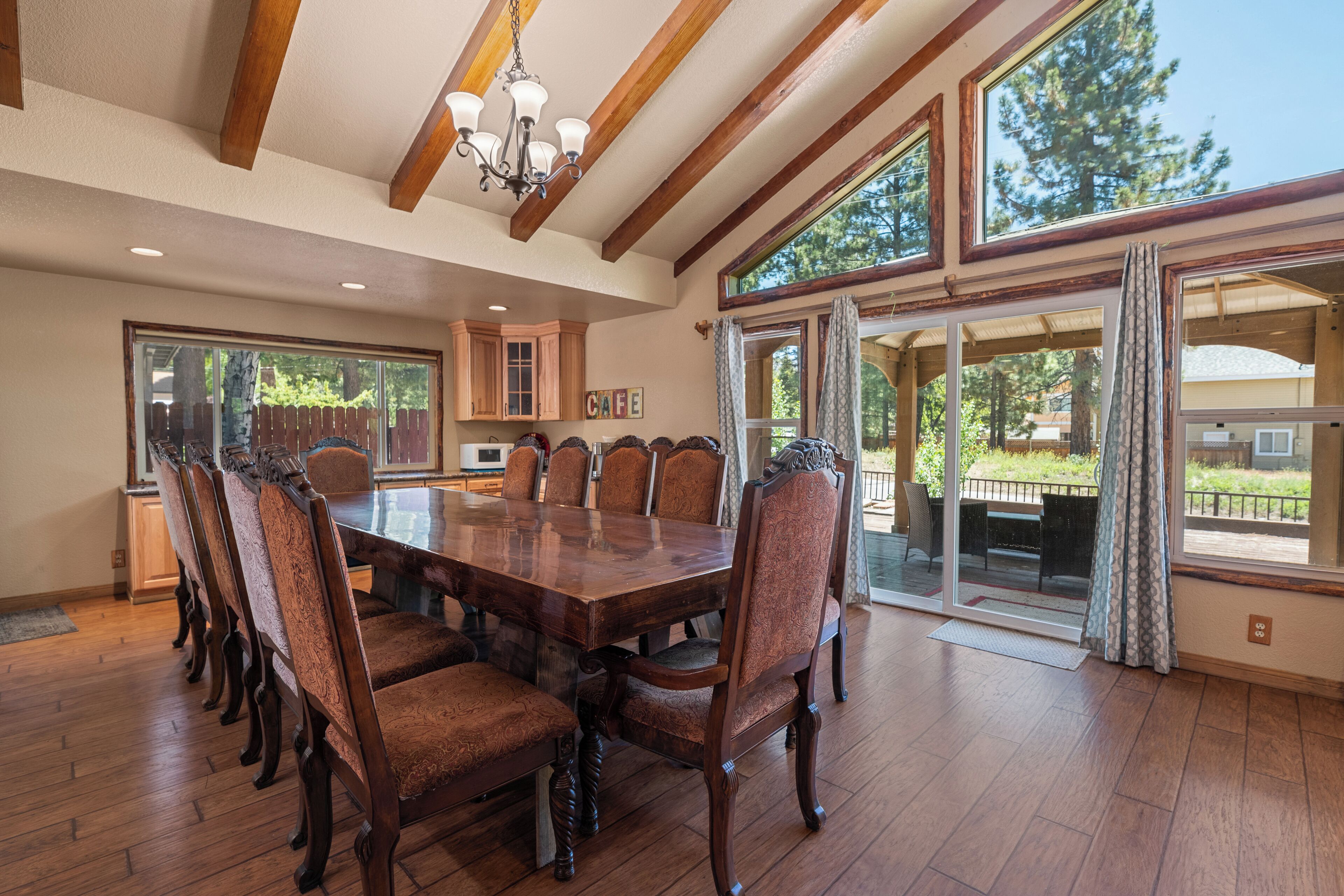 A spacious dining area with a large wooden table and upholstered chairs, a chandelier, and large windows offering views of the outdoors.