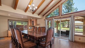A spacious dining area with a large wooden table and upholstered chairs, a chandelier, and large windows offering views of the outdoors. - Moonridge Retreat - 6BR, Walk to Bear Mountain, Foosball Table, BBQ (Big Bear Lake)