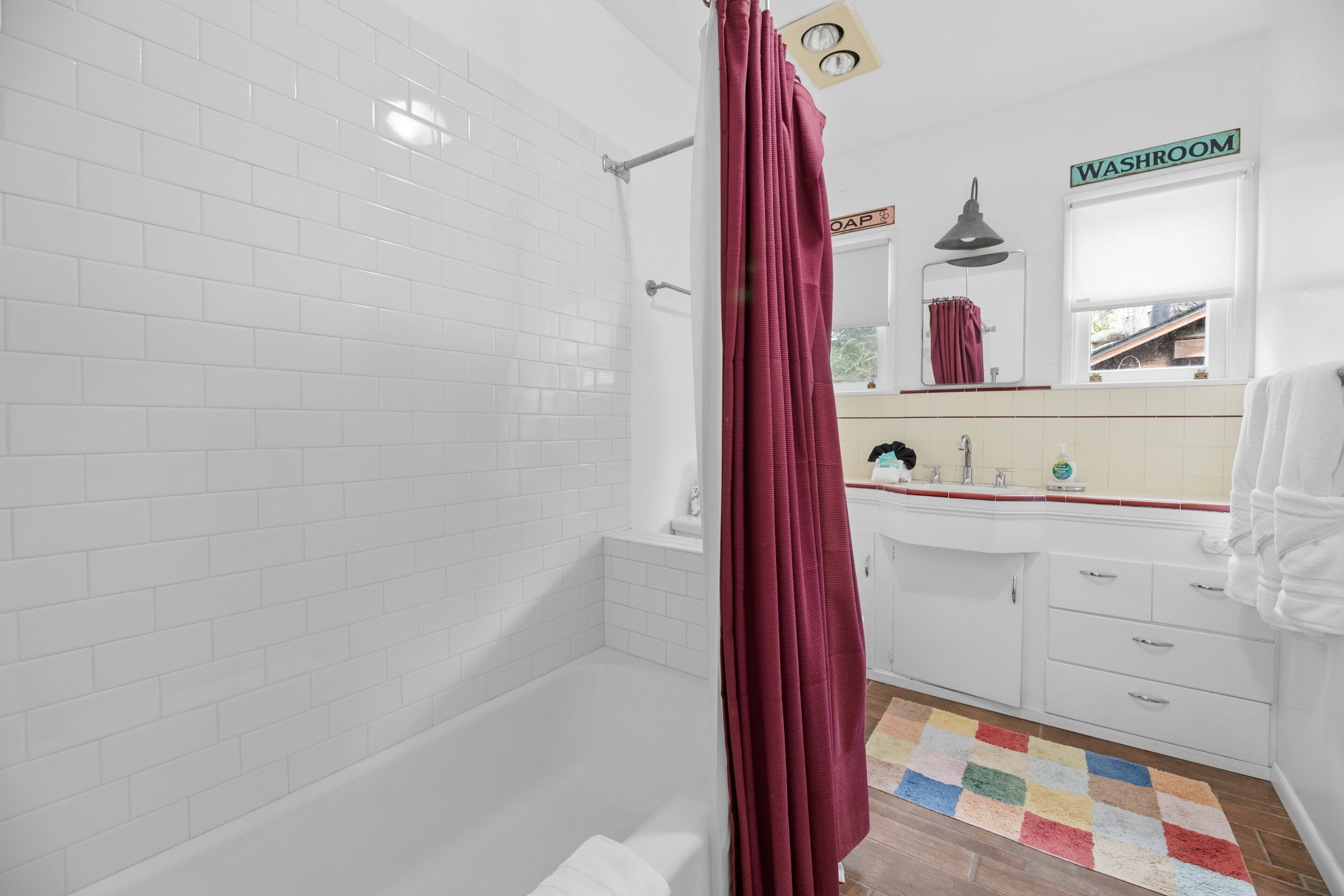 A bathroom with white tiled walls, a bathtub, a shower curtain, a sink with a mirror, and a colorful rug.