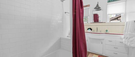 A bathroom with white tiled walls, a bathtub, a shower curtain, a sink with a mirror, and a colorful rug.
