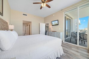 A bedroom with a large bed, a ceiling fan, a television, and a sliding glass door to a balcony.
