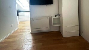 Una habitación con una televisión de pantalla plana montada en la pared, un armario blanco, una mesita de noche pequeña y un radiador.