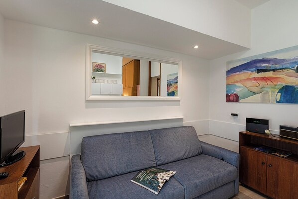 A living room with a grey sofa, a flat-screen TV on a wooden stand, a window with a view of a room with a painting, and a colorful abstract painting on the wall. - Mondial Apartment by Wonderful Italy (Rapallo)