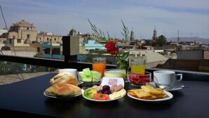 A breakfast table with various food items and drinks, a rose, and a view of a cityscape with buildings and a mosque. - Hotel TerraMistica Monasterio  (Arequipa)