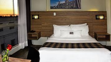 Habitación de hotel con una cama grande, dos mesitas de noche, una ventana con cortinas y vistas a la ciudad.