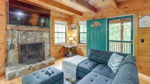 Un interior de cabaña acogedor, con chimenea de piedra, televisor de pantalla plana grande, puerta verde y una zona de estar cómoda con sofá y reposapiés.