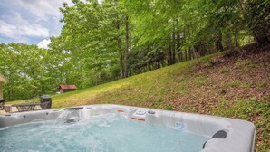 A hot tub in a lush, green outdoor setting with a wooden deck and a small building in the background.