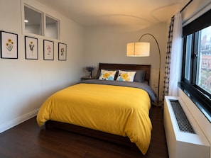 Une chambre à coucher comprenant un lit, une table de chevet, une lampe, une fenêtre avec des rideaux et des reproductions encadrées de motifs botaniques accrochées au mur.