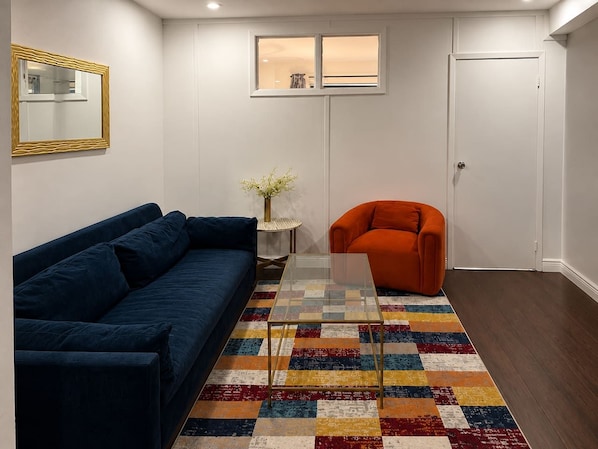 Una sala de estar con un sofá azul, un sillón naranja, una mesa de centro de vidrio y un tapete colorido.