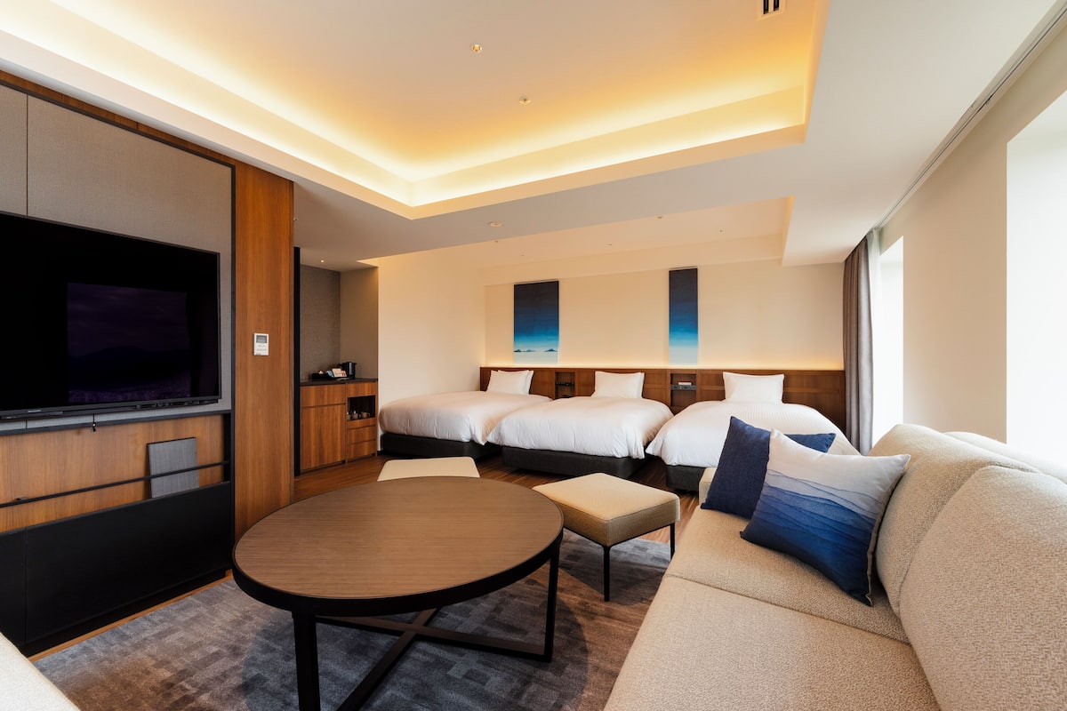 Une chambre d’hôtel moderne dotée d’un grand téléviseur à écran plat, d’une table de salle à manger ronde, d’un canapé et de deux lits avec des têtes de lit assorties.