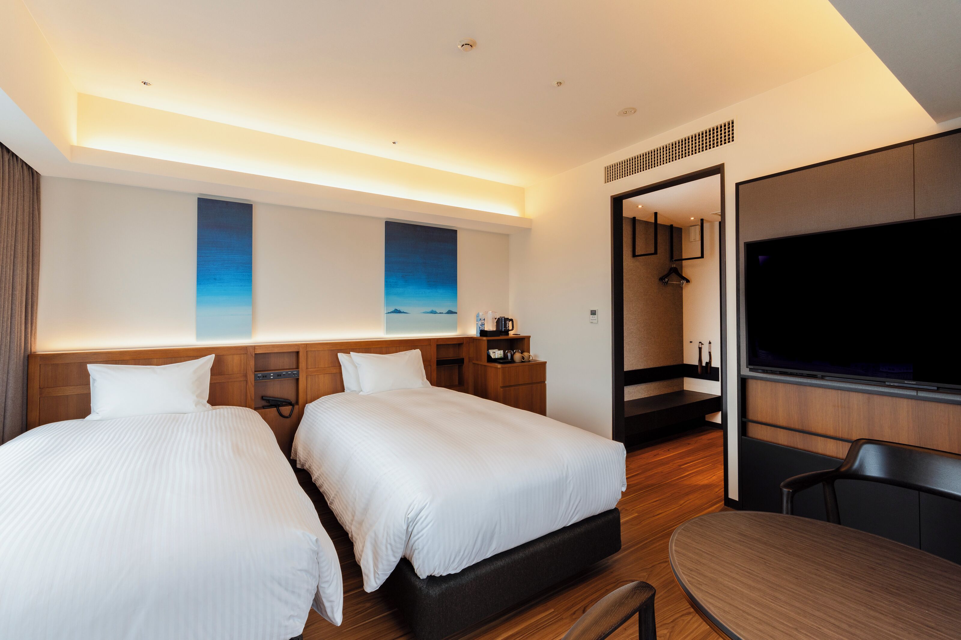 A modern hotel room with two beds, a flat-screen TV, a wooden desk, and a large mirror.