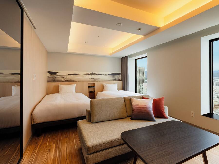A modern hotel room with a large bed, a sofa, a small table, and a city view through a large window.