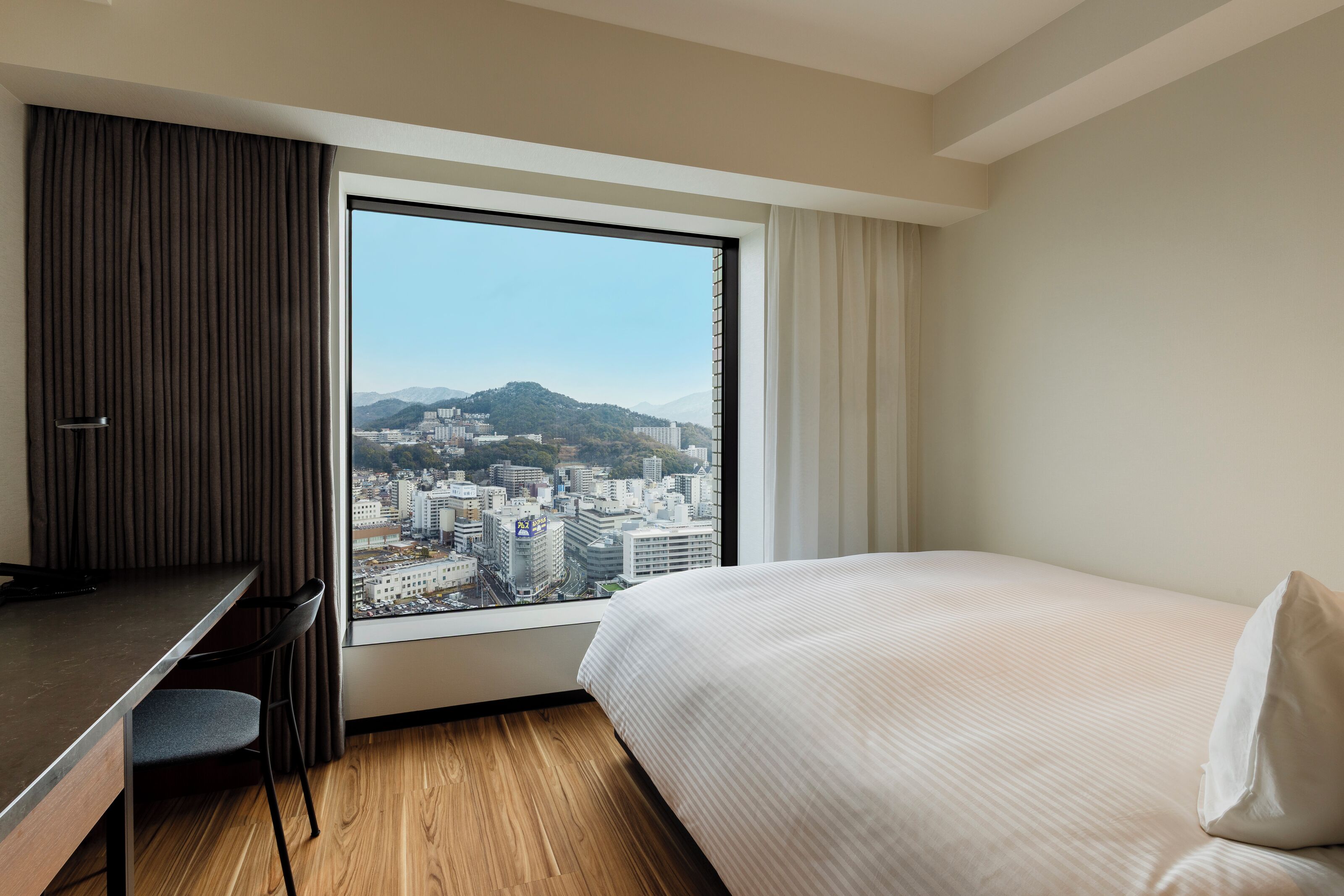 A hotel room with a large window, a bed, a desk, and a chair.