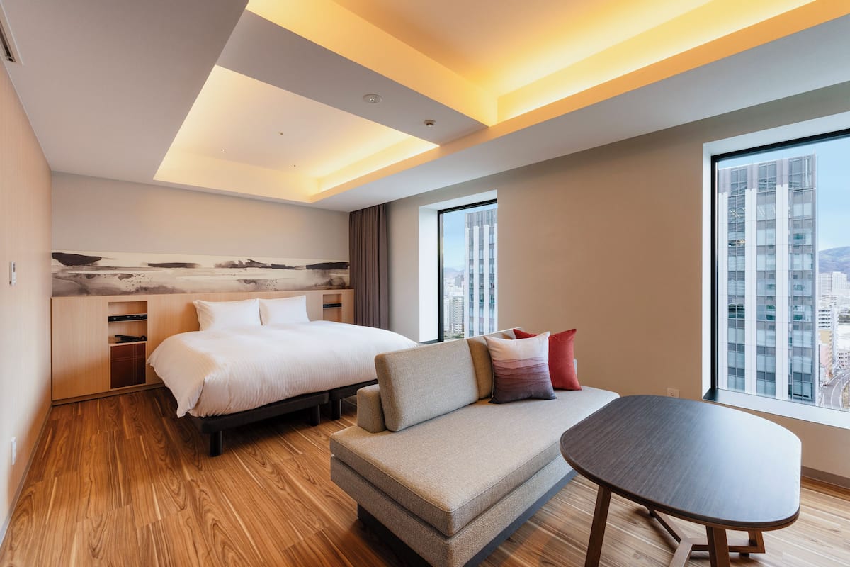Une chambre d’hôtel moderne dotée d’un lit, d’un canapé et d’une table à manger, de grandes fenêtres offrant une vue sur la ville et d’un plancher en bois.