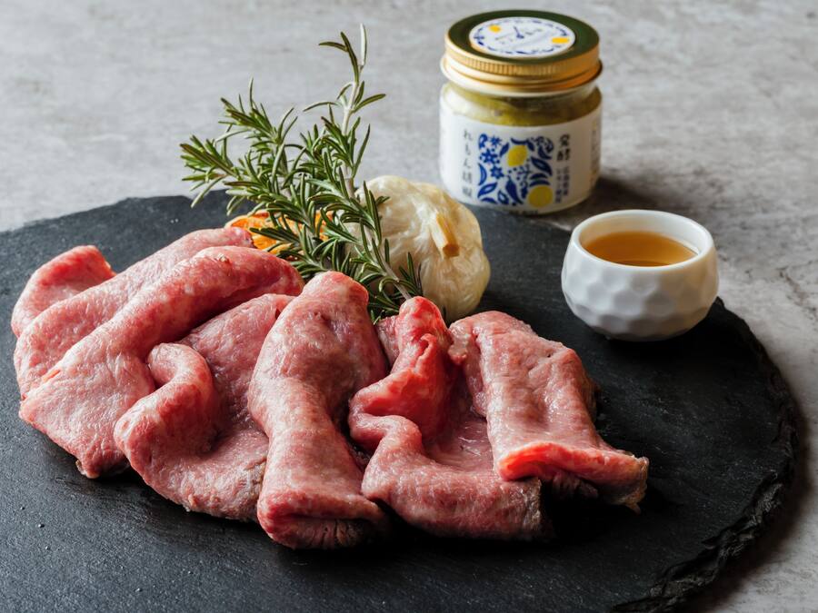Raw meat, a sprig of rosemary, a garlic bulb, a jar of sauce, and a small bowl of sauce on a slate surface.