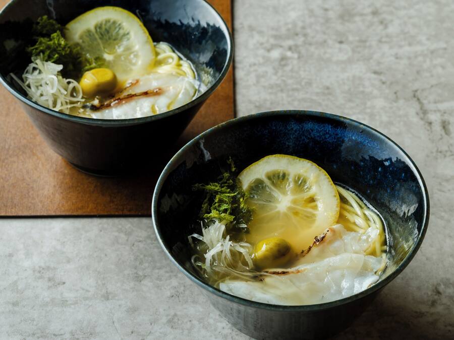 Two bowls of a clear soup with lemon slices and vegetables on a marble surface.