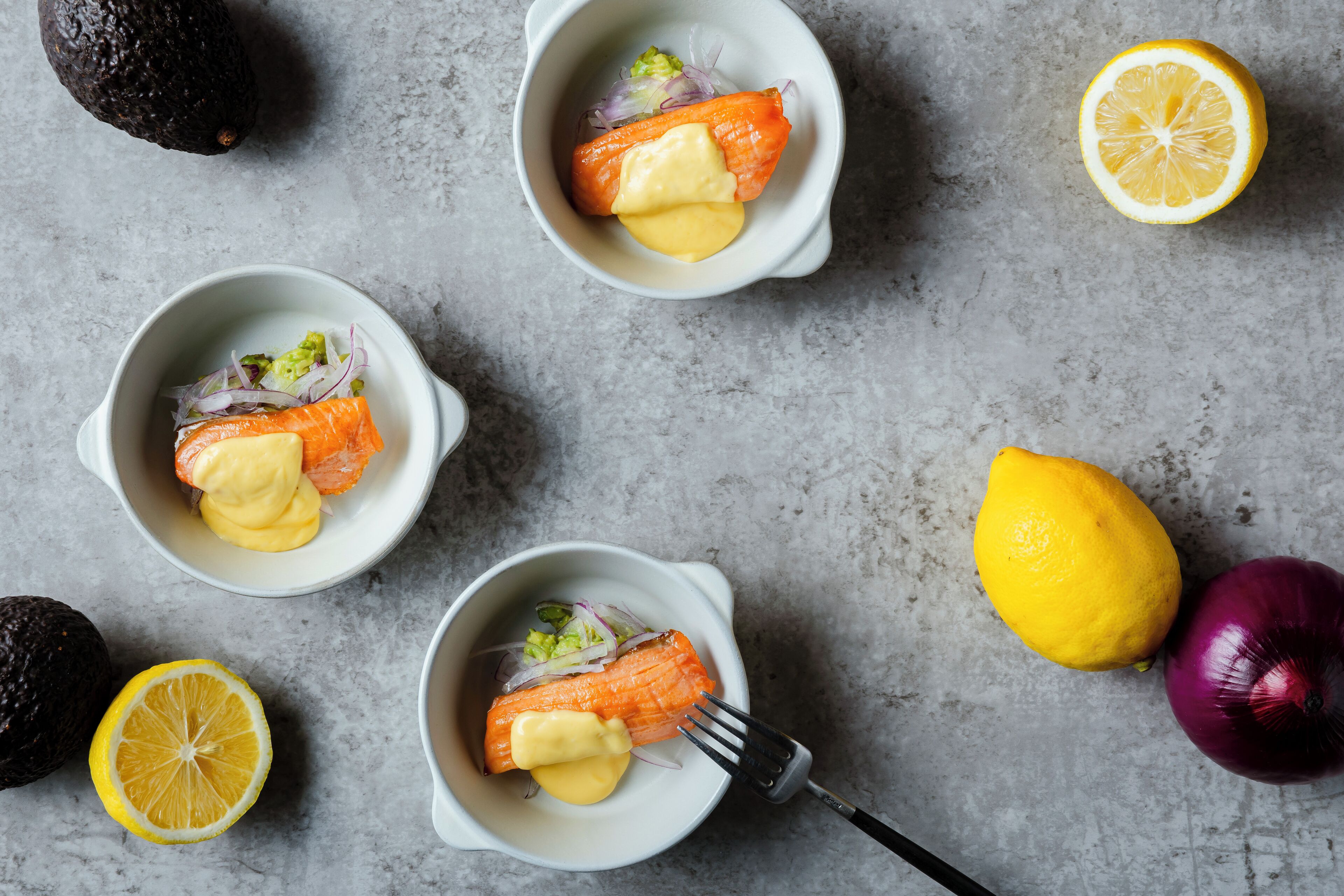 Three bowls of salmon with sauce and vegetables on a gray surface.