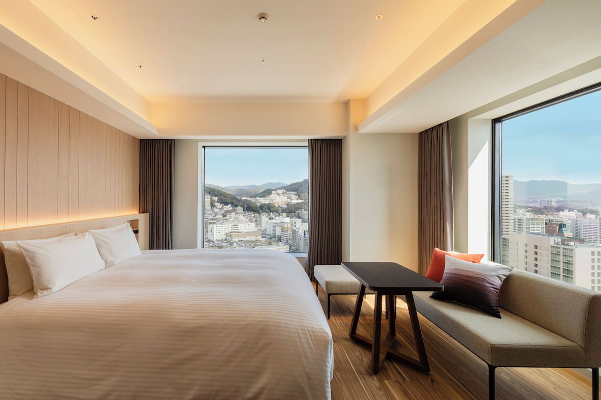 Une chambre d’hôtel moderne, dotée d’un grand lit, d’une petite table, d’un canapé et offrant une vue sur la ville grâce à de grandes fenêtres.