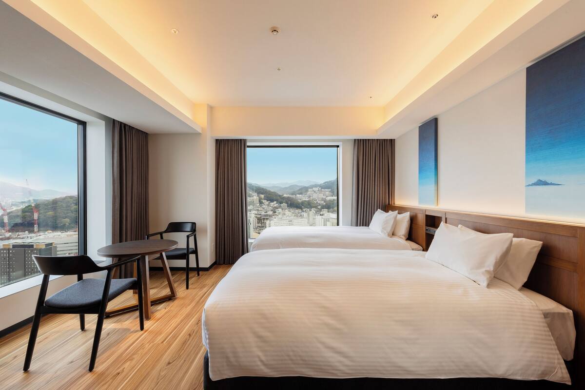 Une chambre d’hôtel avec deux lits, une petite table et une chaise. Une grande fenêtre offre une vue sur la ville.