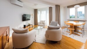 Una sala de estar moderna con un sofá, una mesa de comedor con sillas, un televisor montado en la pared y vistas de la ciudad a través de ventanas amplias.