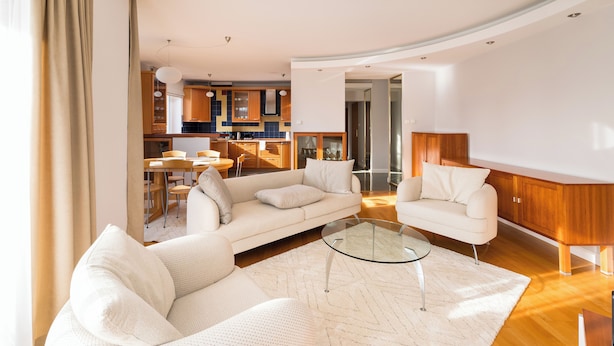Una sala moderna con un sofá, sillones y una mesa de centro de vidrio. Se ve una cocina al fondo con armarios de madera y un comedor.