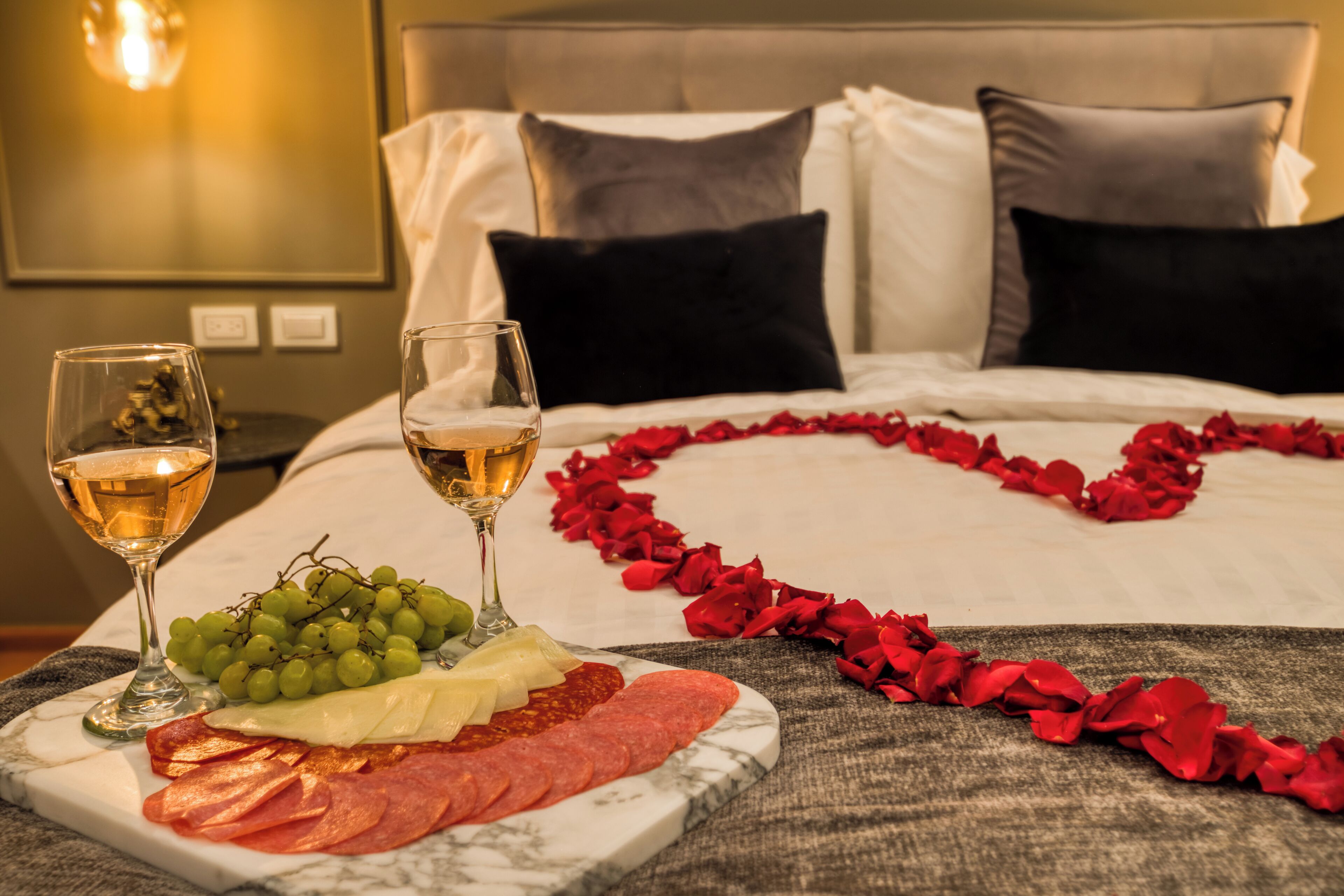 A bed with a heart-shaped arrangement of rose petals, a plate of charcuterie, and two glasses of wine on a marble table.