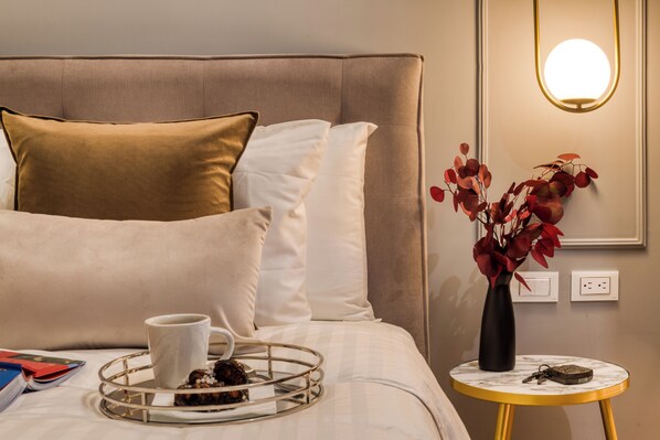 Un lit bien fait, avec un plateau de collations, une tasse et des livres, un vase rempli de fleurs rouges et une petite table ronde surmontée d’une lampe.