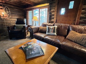 A log cabin interior with a leather sofa, a wooden coffee table, a leather armchair, and a window with blinds.